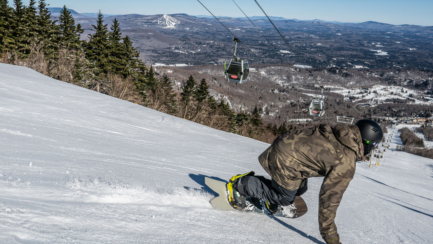 A snowboarder carves a turn on a snowy slope with gondolas and distant mountains